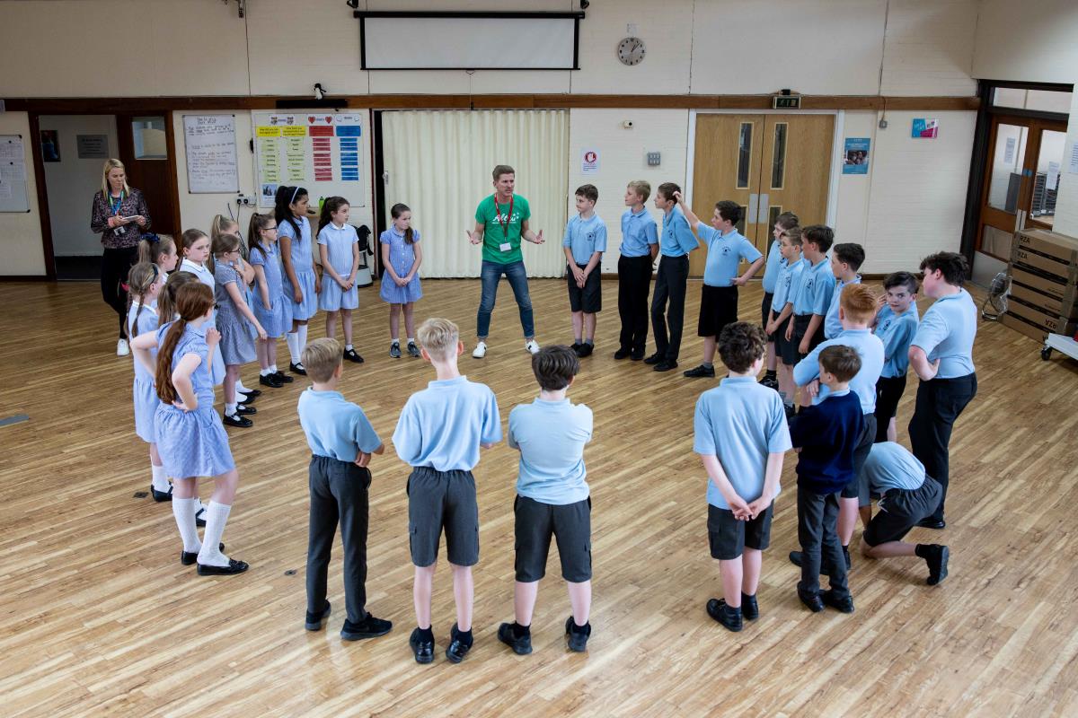 Group of Key stage 2 pupils standing in a circle in school hall with drama workshop leader