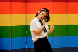 Actor performing as school girl singing along to music track with headphones on in front of a rainbow coloured screen.